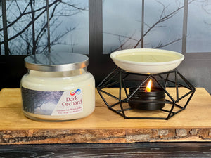 Jar of 'Dark Orchard' candle with a lit wax warmer on a wooden surface. Jar of 'Dark Orchard' candle with a lit wax warmer on a wooden surface.