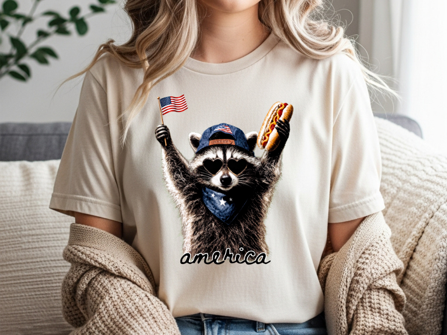 Person wearing a t-shirt with a raccoon graphic holding an American flag and hot dog, sitting on a couch.
