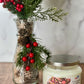 Decorative glass vase with greenery and red berries next to a candle labeled 'Eucalyptus & Apple' on a wooden surface.