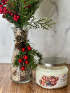 Decorative glass vase with greenery and red berries next to a candle labeled 'Eucalyptus & Apple' on a wooden surface.
