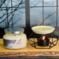 Candle and candle warmer on a wooden surface with a window view of trees and sky.
